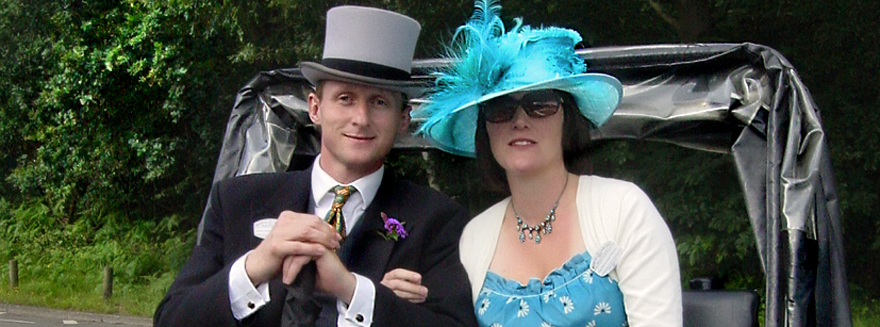 A man in a morning suit and top hat with a woman wearing a blue dress and hat are posing seated on a Bugbugs rickshaw