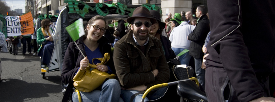 Three people seated on a rickshaw, smiling and waving a flag during an event for the Irish Cultural Centre